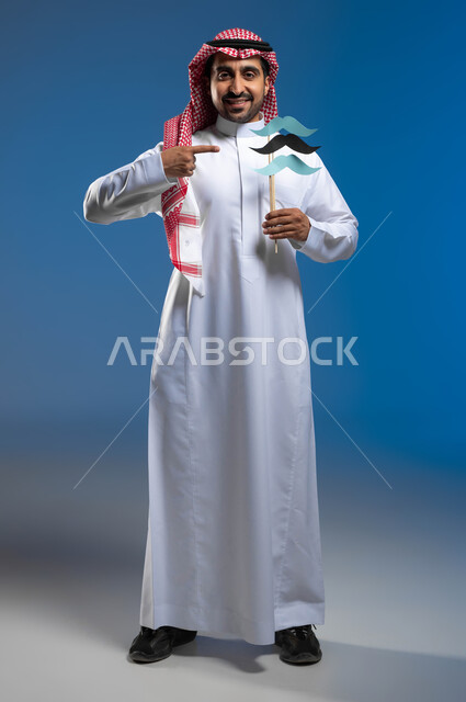 النظر الى الكاميرا بايماءات الفخر والإعتزاز بالنفس، الإهتمام بصحة الرجال، ذكرى اليوم العالمي للرجل 19 نوفمبر، بورتريه لشاب عربي خليجي سعودي يرتدي الثوب التقليدي والشماغ يقوم بالإشارة إلى عود خشبي عليه قصاصات ورقية على شكل شارب، الطول الكامل للجسم، خلفية زرقاء