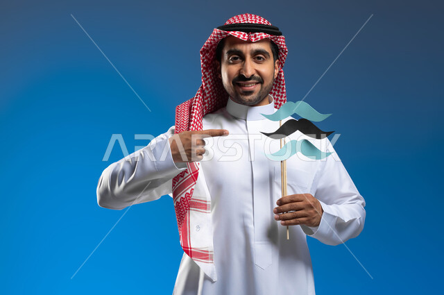 مفهوم الإهتمام بصحة الرجال، النظر الى الكاميرا بايماءات الفخر والإعتزاز بالنفس، ذكرى اليوم العالمي للرجل 19 نوفمبر، بورتريه مقرب لشاب عربي خليجي سعودي يرتدي الثوب التقليدي والشماغ يقوم بالإشارة إلى عود خشبي عليه قصاصات ورقية على شكل شارب، خلفية زرقاء