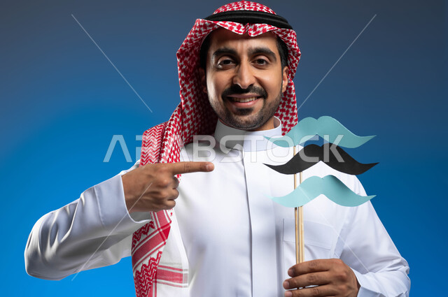 ذكرى اليوم العالمي للرجل 19 نوفمبر، مفهوم الإهتمام بصحة الرجال، النظر الى الكاميرا بايماءات الفخر والإعتزاز بالنفس، بورتريه مقرب لشاب عربي خليجي سعودي يرتدي الثوب التقليدي والشماغ يقوم بالإشارة إلى عود خشبي عليه قصاصات ورقية على شكل شارب، خلفية زرقاء