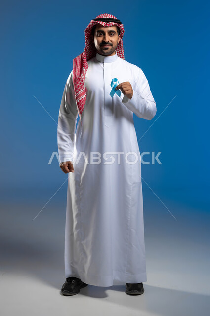 الكشف المبكر عن الأمراض، إتباع الإرشادات الطبية وعدم إهمال الصحة الجسدية، بورتريه لرجل عربي خليجي سعودي يرتدي الثوب التقليدي والشماغ يحمل بيده شريط أزرق للتوعية بسرطان البروستات، صورة الجسم بالطول الكامل، خلفية زرقاء