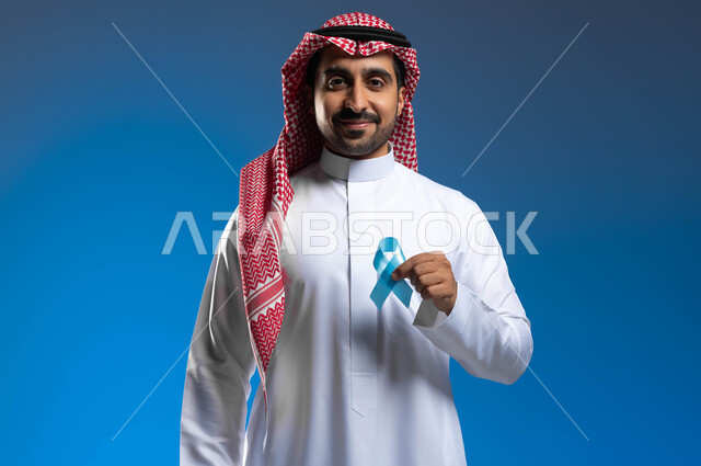 أهمية الكشف المبكر عن الأمراض، إتباع الإرشادات الطبية وعدم إهمال الصحة الجسدية، بورتريه مقرب لرجل عربي خليجي سعودي يرتدي الثوب التقليدي والشماغ يحمل بيده شريط أزرق للتوعية بسرطان البروستات، خلفية زرقاء