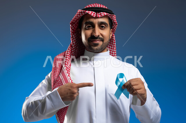 أهمية الكشف المبكر عن السرطانات، حملات التوعية والإرشاد بعدم إهمال الصحة، بورتريه مقرب لرجل عربي خليجي سعودي يرتدي الثوب التقليدي والشماغ يقوم بالإشارة الى الشريط الأزرق للدلالة على سرطان البروستات، خلفية زرقاء
