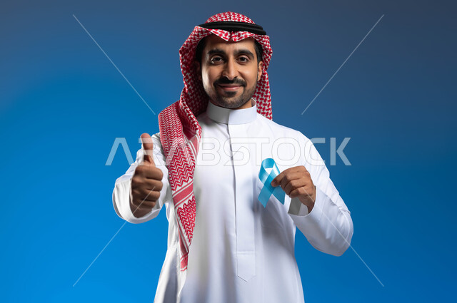 الحفاظ على الصحة الجسدية، رفع اليد بإيماءات الاعجاب والرضا، حملات الإرشاد والتنبيه للكشف المبكر عن سرطان  البروستات، بورتريه مقرب لشاب عربي خليجي سعودي يرتدي الزي التقليدي والشماغ يحمل شريط أزرق للتوعية بالأمراض، خلفية زرقاء