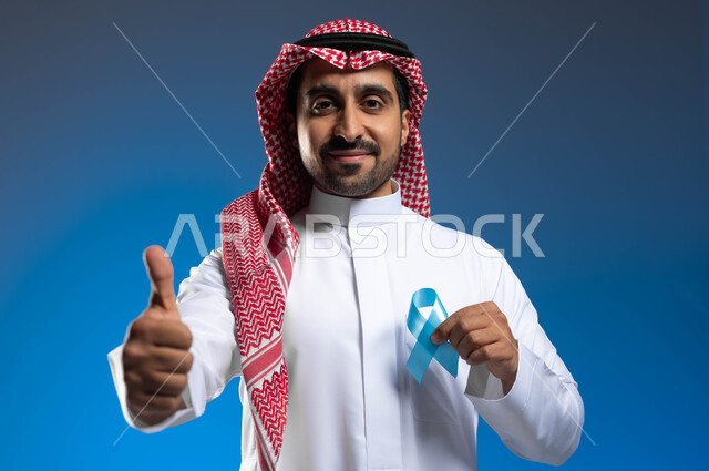 رفع اليد بإيماءات الاعجاب والرضا، الحفاظ على الصحة الجسدية، حملات الإرشاد والتنبيه للكشف المبكر عن سرطان  البروستات، بورتريه مقرب لشاب عربي خليجي سعودي يرتدي الزي التقليدي والشماغ يحمل شريط أزرق للتوعية بالأمراض، خلفية زرقاء