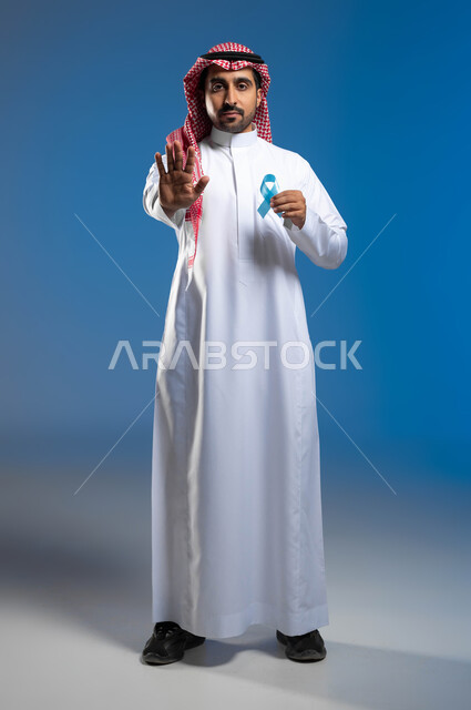 حملات التوعية والإرشاد للكشف المبكر عن الأمراض، بورتريه لرجل عربي خليجي سعودي يرتدي الزي التقليدي يحمل شريط أزرق للتوعية بسرطان البروستات، الوقوف باستقامة بإيماءات الحزم والجدية، رفع الكف مفتوحة للتحذير من تجاهل الكشف عن المرض، صورة لكامل الجسم، خلفية زرقاء