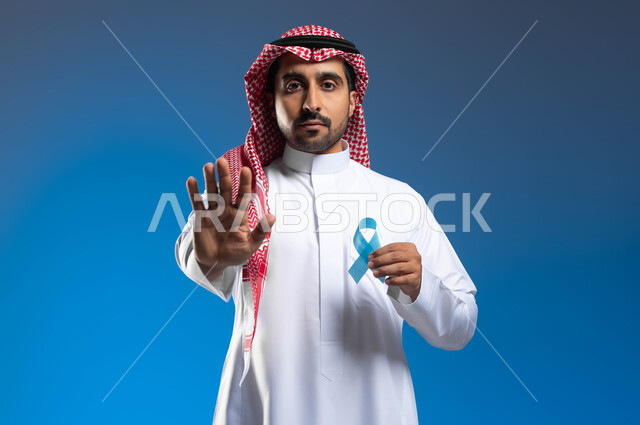 رفع الكف مفتوحة للتحذير من تجاهل الكشف عن المرض، حملات التوعية والإرشاد للكشف المبكر عن الأمراض، بورتريه لرجل عربي خليجي سعودي يرتدي الزي التقليدي يحمل شريط أزرق للتوعية بسرطان البروستات، الوقوف باستقامة بإيماءات الحزم والجدية، خلفية زرقاء