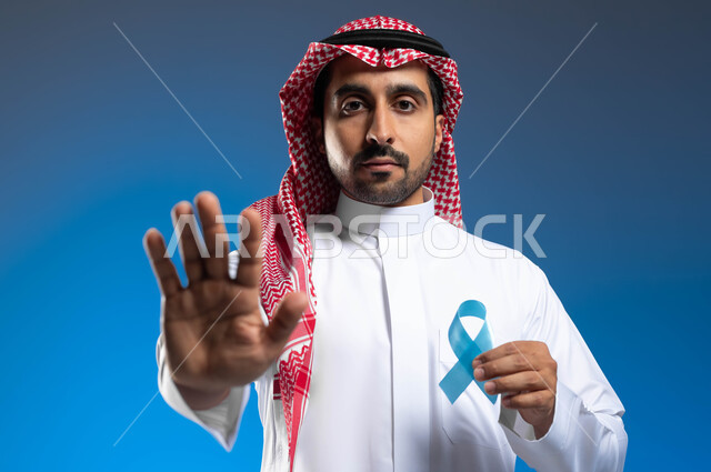 شعار الكشف المبكر عن الأمراض، رفع الكف مفتوحة للتحذير من إهمال الصحة الجسدية، بورتريه مقرب لرجل عربي خليجي سعودي يرتدي الزي التقليدي يحمل شريط أزرق للتوعية بسرطان البروستات، الوقوف باستقامة بإيماءات الحزم والجدية، خلفية زرقاء