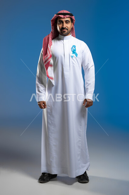 الوقوف باستقامة والنظر إلى الكاميرا بإيماءات الحزم والجدية، إتباع الأساليب الوقائية عن طريق الكشف المبكر عن سرطان البروستات، بورتريه لشاب عربي خليجي سعودي يرتدي الزي التقليدي والشماغ يضع على صدره شريط أزرق للتوعية بالأمراض، صورة لكامل الجسم، خلفية زرقاء