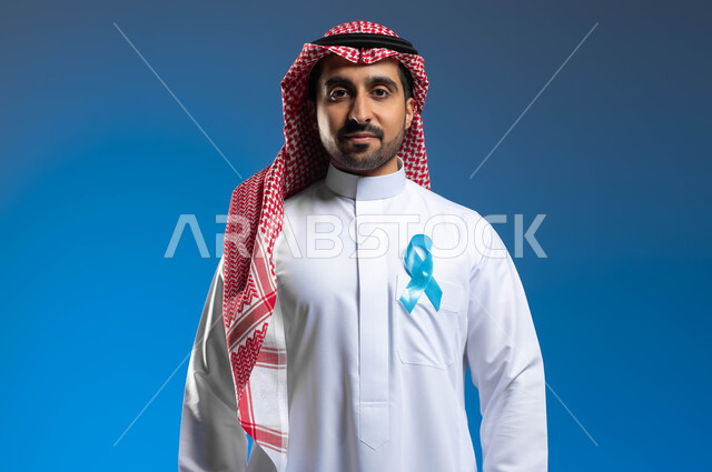 إتباع الأساليب الوقائية والحفاظ على الصحة الجسدية، الكشف المبكر عن سرطان البروستات، الوقوف باستقامة والنظر إلى الكاميرا بإيماءات الحزم والجدية، بورتريه لشاب عربي خليجي سعودي يرتدي الزي التقليدي والشماغ يضع على صدره شريط أزرق للتوعية بالأمراض، خلفية زرقاء