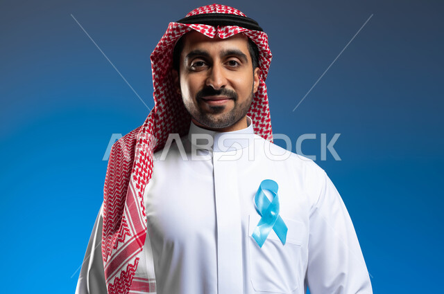 الكشف المبكر عن سرطان البروستات، إتباع الأساليب الوقائية والحفاظ على الصحة الجسدية، الوقوف باستقامة والنظر إلى الكاميرا، بورتريه مقرب لشاب عربي خليجي سعودي يرتدي الزي التقليدي والشماغ يضع على صدره شريط أزرق للتوعية بالأمراض، خلفية زرقاء
