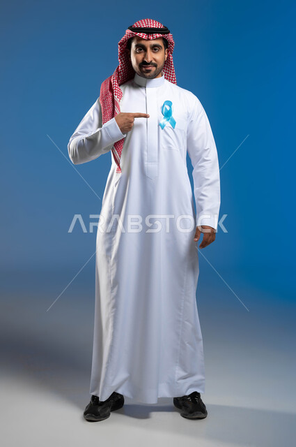 الحفاظ على الصحة الجسدية وإتباع الأساليب الوقائية، الوقوف باستقامة والنظر إلى الكاميرا بإيماءات الرضا، الكشف المبكر عن سرطان البروستات، بورتريه لشاب عربي خليجي سعودي يرتدي الزي التقليدي يشير بإصبعه إلى شريط أزرق على صدره، صورة لكامل الجسم، خلفية زرقاء