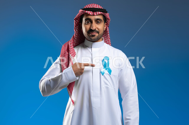 إتباع الأساليب الوقائية والحفاظ على الصحة الجسدية، الوقوف باستقامة والنظر إلى الكاميرا بإيماءات التفاؤل والرضا، الكشف المبكر عن سرطان البروستات، بورتريه مقرب لشاب عربي خليجي سعودي يرتدي الثوب التقليدي والشماغ يشير بإصبعه إلى شريط أزرق على صدره، خلفية زرقاء