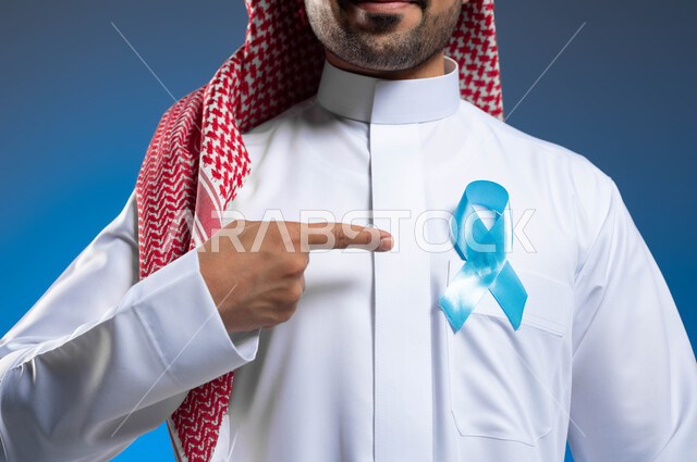 الوقاية من الأمراض عبر الكشف المبكر عن سرطان البروستات، الحفاظ على الصحة الجسدية، بورتريه مقرب لشاب عربي خليجي سعودي يرتدي الشماغ والثوب التقليدي يشير بإصبعه إلى شريط أزرق على صدره، خلفية زرقاء
