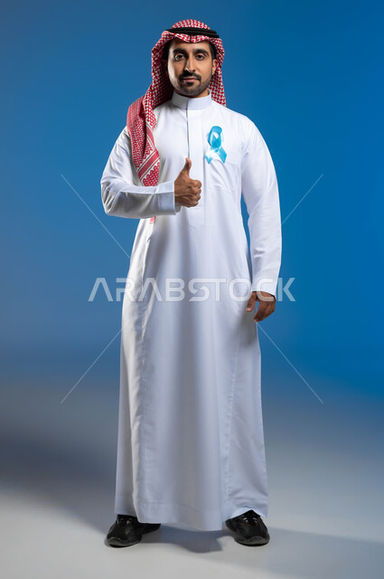 الكشف المبكر عن سرطان البروستات، حملات التوعية والتنبيه بعدم إهمال الصحة الجسدية، رفع اليد بإيماءات الاعجاب والرضا، بورتريه لشاب عربي خليجي سعودي يرتدي الزي التقليدي يضع على صدره شريط أزرق للتوعية بالأمراض، صورة لكامل الجسم، خلفية زرقاء