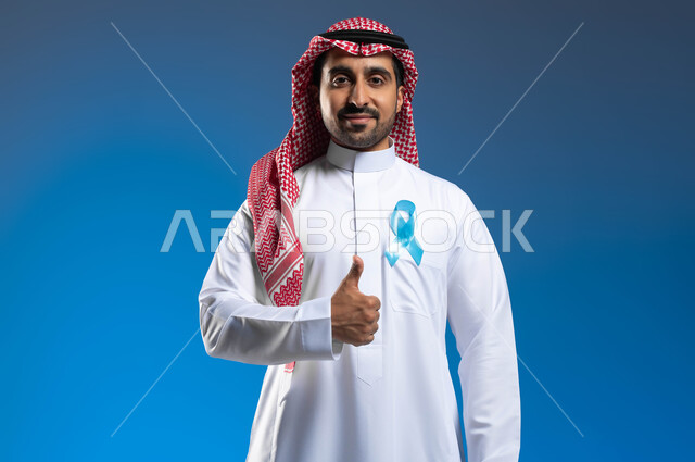 حملات الإرشاد والتنبيه للكشف المبكر عن سرطان  البروستات، الوقوف باستقامة ورفع اليد بإيماءات الاعجاب والرضا، بورتريه  لشاب عربي خليجي سعودي يرتدي الزي التقليدي يضع على صدره شريط أزرق للتوعية بالأمراض، الحفاظ على الصحة الجسدية، خلفية زرقاء