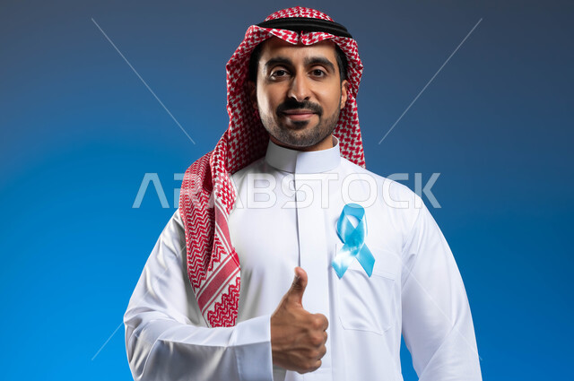 الحفاظ على الصحة الجسدية، حملات الإرشاد والتنبيه للكشف المبكر عن سرطان  البروستات، الوقوف باستقامة ورفع اليد بإيماءات الاعجاب والرضا، بورتريه مقرب لشاب عربي خليجي سعودي يرتدي الزي التقليدي يضع على صدره شريط أزرق للتوعية بالأمراض، خلفية زرقاء