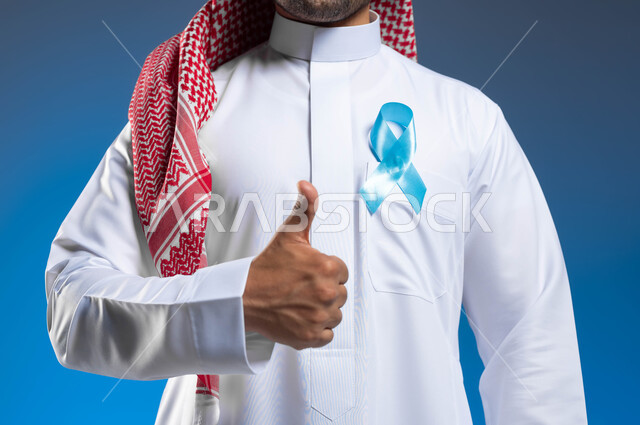 Following preventive methods through early detection of prostate cancer, maintaining physical health, a close-up portrait of a young Saudi Arabian Gulf man wearing a shemagh and traditional dress with a blue ribbon on his chest to raise awareness about diseases, hand gestures indicating admiration and satisfaction, blue background