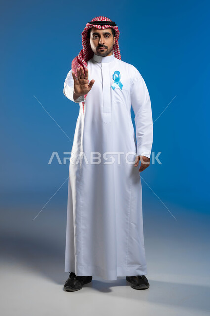 حملات التوعية والإرشاد بعدم إهمال الصحة الجسدية، الوقوف باستقامة والنظر إلى الكاميرا ورفع اليد بإيماءات التحذير والتنبيه، بورتريه لشاب عربي خليجي سعودي يرتدي الزي التقليدي يضع على صدره شعار الكشف المبكر عن سرطان البروستات، صورة لكامل الجسم، خلفية زرقاء