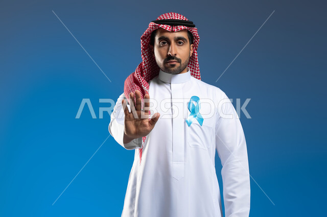 رفع اليد بإشارات التحذير والتنبيه، الوقوف باستقامة والنظر إلى الكاميرا بإيماءات الحزم والجدية، حملات التوعية بعدم إهمال الصحة الجسدية، بورتريه لشاب عربي خليجي سعودي يرتدي الثوب التقليدي والشماغ يضع على صدره شعار الكشف المبكر عن سرطان البروستات، خلفية زرقاء