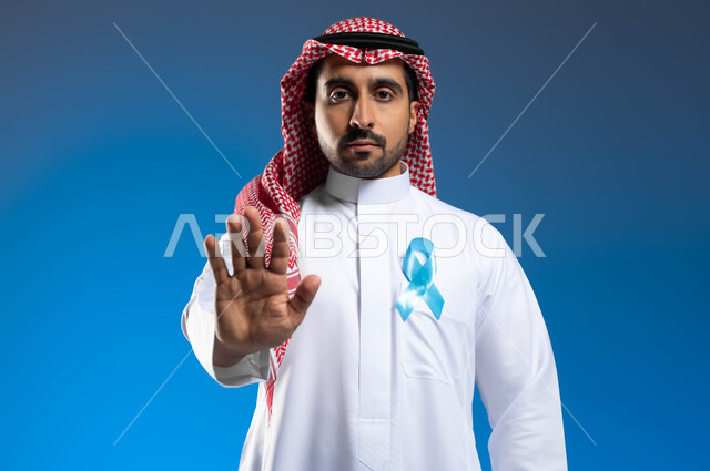 الوقوف باستقامة بإيماءات الحزم والجدية، رفع اليد بإشارات التحذير والتنبيه، حملات التوعية والإرشاد بعدم إهمال الصحة الجسدية، بورتريه مقرب لشاب عربي خليجي سعودي يرتدي الثوب التقليدي والشماغ يضع على صدره شعار الكشف المبكر عن سرطان البروستات، خلفية زرقاء