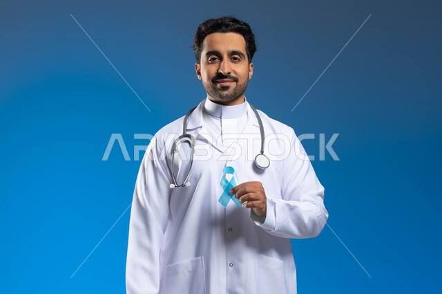 تقديم الخدمات الصحية للمرضى، حملات التوعية والتنبيه، القيام بإجراءات الوقاية من السرطانات، بورتريه مقرب لطبيب عربي خليجي سعودي يرتدي المعطف الطبي والسماعات الطبية يحمل بيده شعار الكشف المبكر عن سرطان البروستات، خلفية زرقاء