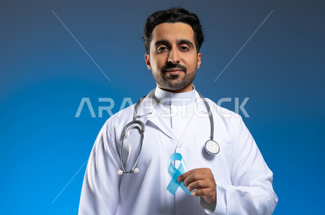 Awareness and warning campaigns, taking cancer prevention measures, providing health services to patients, a close-up portrait of a Saudi Gulf Arab doctor wearing a medical coat and stethoscopes, holding the slogan for early detection of prostate cancer in his hand, blue background.