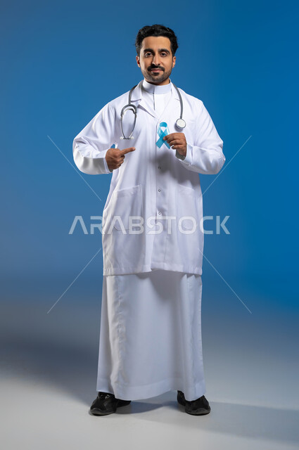 مفهوم الطب والرعاية الصحية، الكشف المبكر عن الأمراض وإتباع أساليب الوقاية، بورتريه لطبيب عربي خليجي سعودي يرتدي المعطف الطبي والسماعات الطبية يقوم بالإشارة بإصبعه السبابة الى شعار التوعية بسرطان البروستات، الطول الكامل للجسم، خلفية زرقاء