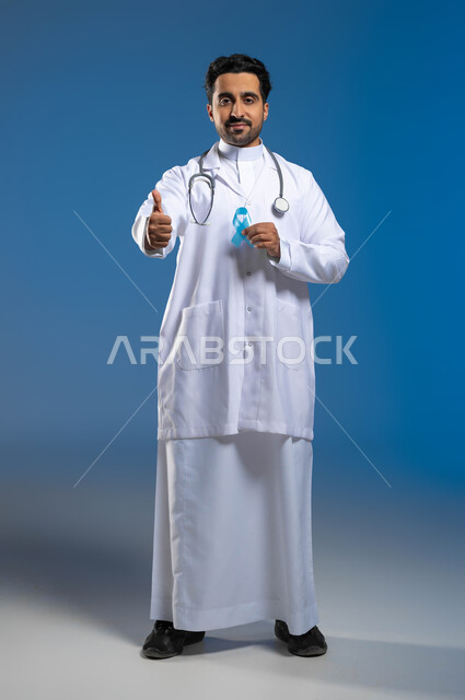 تعابير الإعجاب والموافقة على الكشف المبكر، مفهوم العمل في المراكز الصحية والمستشفيات، بورتريه لطبيب عربي خليجي سعودي يرتدي المعطف الطبي والسماعات الطبية يحمل بيده شريط أزرق للتوعية بسرطان البروستات، الطول الكامل للجسم، خلفية زرقاء