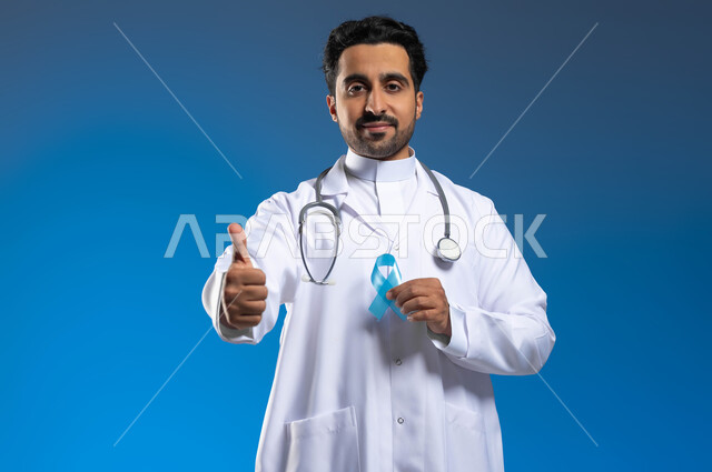 مفهوم العمل في المراكز الصحية والمستشفيات، ايماءات الإعجاب والموافقة على الكشف المبكر، بورتريه مقرب لطبيب عربي خليجي سعودي يرتدي المعطف الطبي والسماعات الطبية يحمل بيده شريط أزرق للتوعية بسرطان البروستات، خلفية زرقاء