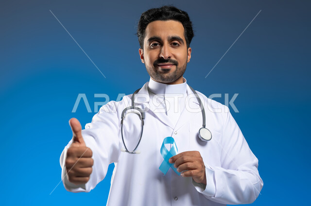 ايماءات الإعجاب والموافقة على الكشف المبكر، مفهوم العمل في المراكز الصحية والمستشفيات، بورتريه مقرب لطبيب عربي خليجي سعودي يرتدي المعطف الطبي والسماعات الطبية يحمل بيده شريط أزرق للتوعية بسرطان البروستات، خلفية زرقاء