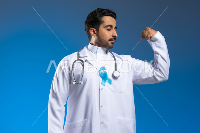 اتباع اساليب الوقاية والكشف المبكر عن الأمراض، العمل في المراكز الصحية والمستشفيات، حملات التوعية بسرطان البروستات، بورتريه مقرب لطبيب عربي خليجي سعودي يرتدي المعطف الطبي والسماعات الطبية يرفع يده بايماءات القوة العضلية والصحة الجسدية، خلفية زرقاء