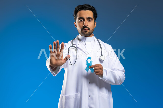 رفع الكف مفتوحة للتحذير من تجاهل الكشف عن المرض، مفهوم العمل في القطاع الصحي، بورتريه مقرب لطبيب عربي خليجي سعودي يرتدي المعطف الطبي يحمل شريط ازرق للتوعيه بسرطان البروستات، ايماءات التوقف عن إهمال  الصحه الجسديه، خلفية زرقاء