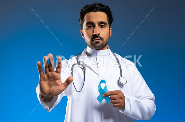 مفهوم العمل في القطاع الصحي، بورتريه مقرب لطبيب عربي خليجي سعودي يرتدي المعطف الطبي يحمل شريط ازرق للتوعيه بسرطان البروستات، رفع الكف مفتوحة للتحذير من تجاهل الكشف عن المرض،  ايماءات التوقف عن إهمال  الصحه الجسديه، خلفية زرقاء