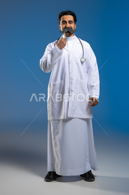Celebration of International Men’s Day, November 19, concept of medicine and health care, work in the health sector, portrait of a Saudi Gulf Arab doctor wearing a medical coat and stethoscopes, holding a wooden stick in his hand with a paper clip in the shape of a mustache, full length of the body, blue background