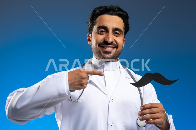 الإشارة الى الورقة بإيماءات الفرح والفخر، سنوية اليوم العالمي للرجل، مفهوم الطب والرعاية الصحية، بورتريه مقرب لطبيب عربي خليجي سعودي يرتدي المعطف الطبي والسماعات الطبية يحمل بيده عود خشبي مع قصاصة ورقية على شكل شارب، خلفية زرقاء