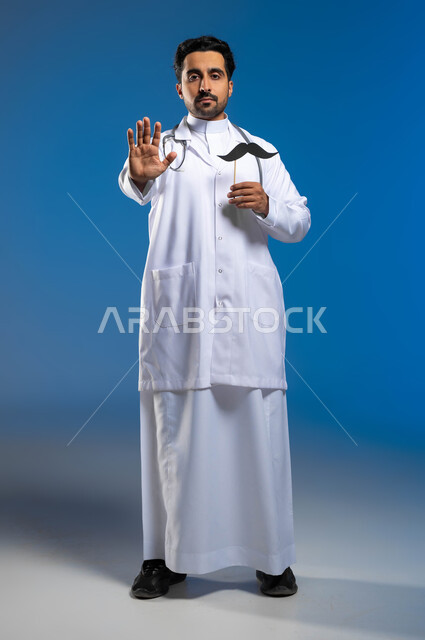 سنوية اليوم العالمي للرجل، ايماءات يدين تدل على الحذر وترك مسافة للحماية من العدوى، مفهوم الطب والرعاية الصحية، بورتريه لطبيب عربي خليجي سعودي يرتدي المعطف الطبي والسماعات الطبية يحمل بيده عود خشبي مع قصاصة ورقية على شكل شارب، الطول الكامل للجسم، خلفية زرقاء