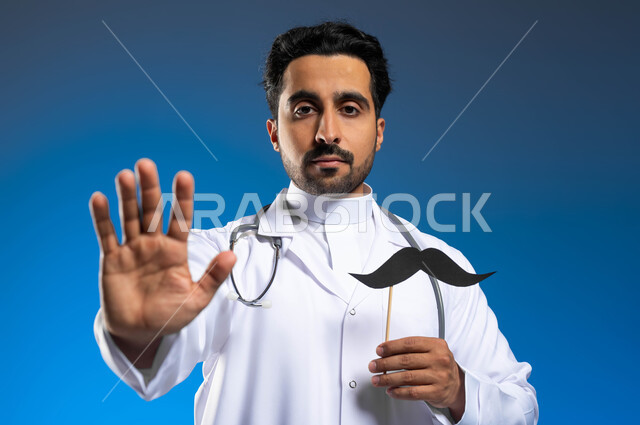 مفهوم الطب والرعاية الصحية، ايماءات يدين تدل على الحذر وترك مسافة أمان للحماية من العدوى، بورتريه مقرب لطبيب عربي خليجي سعودي يرتدي المعطف الطبي والسماعات الطبية يحمل بيده عود خشبي مع قصاصة ورقية على شكل شارب، سنوية اليوم العالمي للرجل، خلفية زرقاء