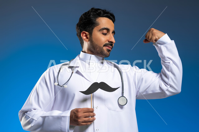 العمل في القطاع الصحي ومفهوم مهنة الطب، ذكرى اليوم العالمي للرجل 19 نوفمبر، الحفاظ على الصحة الجسدية والقوة العضلية، بورتريه مقرب لطبيب عربي خليجي سعودي يرتدي المعطف الطبي والسماعات الطبية يحمل بيده عود خشبي مع قصاصة ورقية على شكل شارب، خلفية زرقاء
