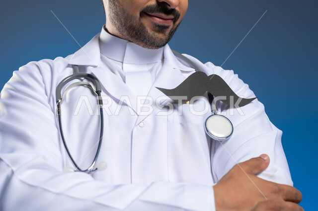 The medical profession and work in the health sector, a close-up portrait of a Saudi Arabian Gulf doctor wearing a medical coat and stethoscopes, standing straight with crossed hands, International Men’s Day, November 19, looking at a piece of paper in the shape of a mustache with proud gestures, blue background