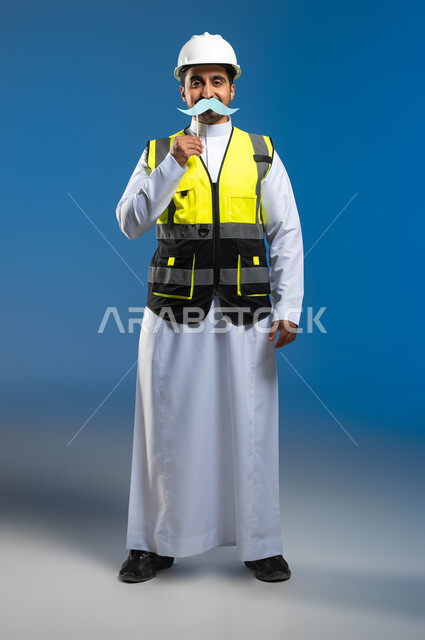 الإشراف وتدقيق الأعمال الهندسية، اليوم العالمي للرجل 19 نوفمبر، مفهوم الهندسة المعمارية والإنشاءات، بورتريه لمهندس عربي خليجي سعودي يرتدي السترة وخوذة الحماية ويمسك عود خشبي مع قصاصة ورقية على شكل شارب، الطول الكامل للجسم، خلفية زرقاء