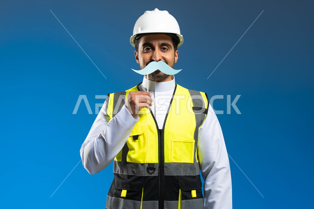 International Men's Day, November 19, concept of architecture and construction, close-up portrait of a Saudi Gulf Arab engineer wearing a jacket and a protective helmet and holding a wooden stick with a clip of paper in the shape of a mustache, supervision and auditing of engineering works, blue background