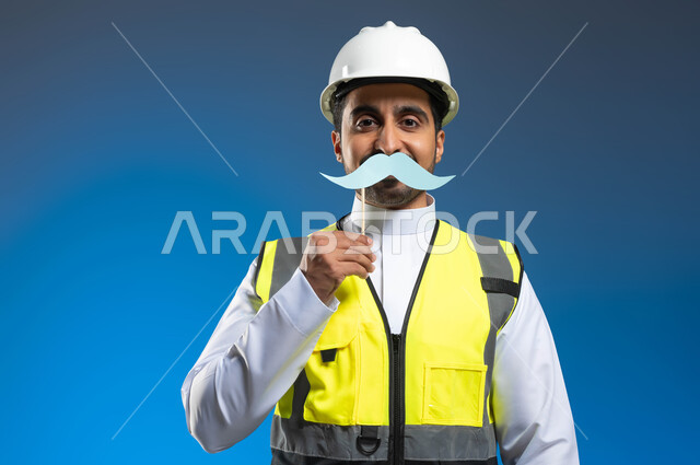 مفهوم الهندسة المعمارية والإنشاءات، اليوم العالمي للرجل 19 نوفمبر، بورتريه مقرب لمهندس عربي خليجي سعودي يرتدي السترة وخوذة الحماية ويمسك عود خشبي مع قصاصة ورقية على شكل شارب، الإشراف وتدقيق الأعمال الهندسية، خلفية زرقاء