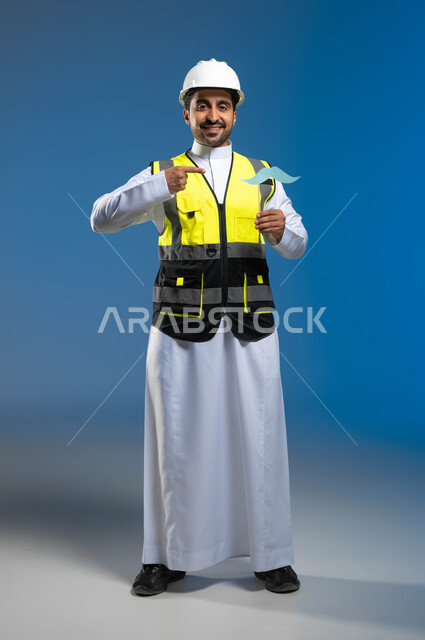 المهن والوظائف الهندسية السعودية، الاحتفال باليوم العالمي للرجل 19 نوفمبر، بورتريه لمهندس عربي خليجي سعودي يرتدي السترة وخوذة الحماية ينظر الى الكاميرا بتعابير السرور، الإشارة باصبع السبابة الى عود خشبي مع قصاصة على شكل شارب، صورة الجسم بالكامل، خلفية زرقاء