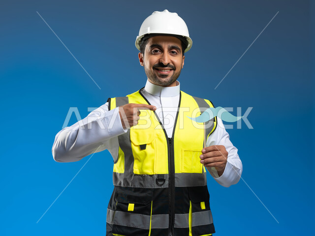 الإشارة باصبع السبابة الى عود خشبي مع قصاصة ورقية على شكل شارب، الوظائف الهندسية في الشركات السعودية، الاحتفال باليوم العالمي للرجل 19 نوفمبر، بورتريه مقرب لمهندس عربي خليجي سعودي يرتدي السترة وخوذة الحماية ينظر الى الكاميرا بتعابير السرور، خلفية زرقاء