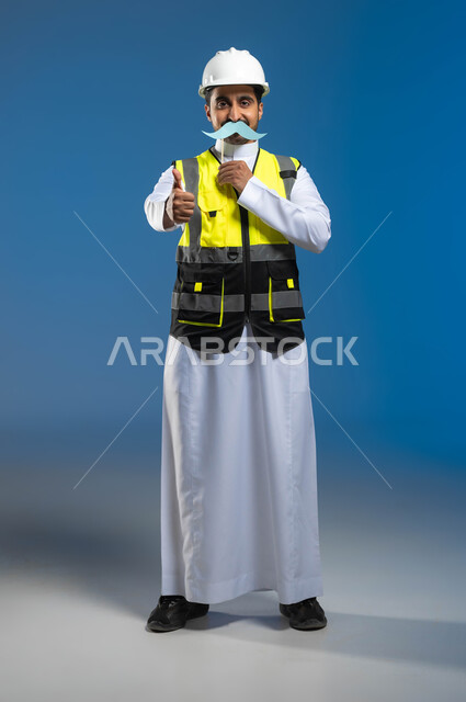 Gestures of approval and admiration for something, commemorating International Men’s Day November 19, portrait of a Saudi Gulf Arab engineer wearing a jacket and a protective helmet and holding a wooden stick with a clip of paper in the shape of a mustache, engineering professions and jobs in Saudi Arabia, full length of the body, blue background