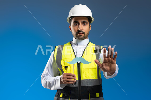 المهن والوظائف الهندسية في السعودية، ايماءات الحذر والابتعاد وترك مسافة، بورتريه مقرب لمهندس عربي خليجي سعودي يرتدي السترة وخوذة الحماية ويمسك عود خشبي مع قصاصة ورقية على شكل شارب، اليوم العالمي للرجل 19 نوفمبر، خلفية زرقاء