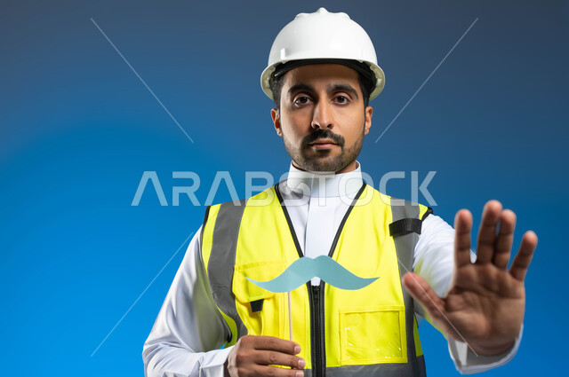 ايماءات الحذر والابتعاد وترك مسافة، المهن والوظائف الهندسية في السعودية، بورتريه مقرب لمهندس عربي خليجي سعودي يرتدي السترة وخوذة الحماية ويمسك عود خشبي مع قصاصة ورقية على شكل شارب، اليوم العالمي للرجل 19 نوفمبر، خلفية زرقاء