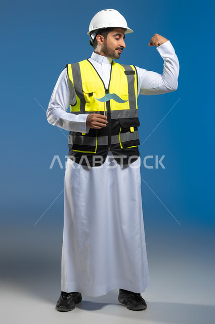مفهوم العمل في قطاع الهندسة المعمارية، الإحتفال السنوي باليوم العالمي للرجل، بورتريه لمهندس عربي خليجي سعودي يرتدي السترة وخوذة الحماية ويمسك عود خشبي مع قصاصة ورقية على شكل شارب، ايماءات الثقة والحفاظ على اللياقة البدنية، الطول الكامل للجسم، خلفية زرقاء