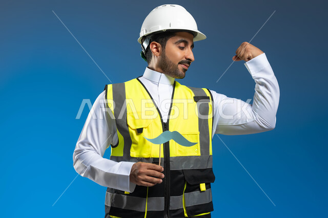 Gestures of confidence and maintaining physical fitness, the concept of work in the architecture sector, a close-up portrait of a Saudi Gulf Arab engineer wearing a jacket and a protective helmet and holding a wooden stick with a paper clip in the shape of a mustache, the annual celebration of International Men’s Day, blue background