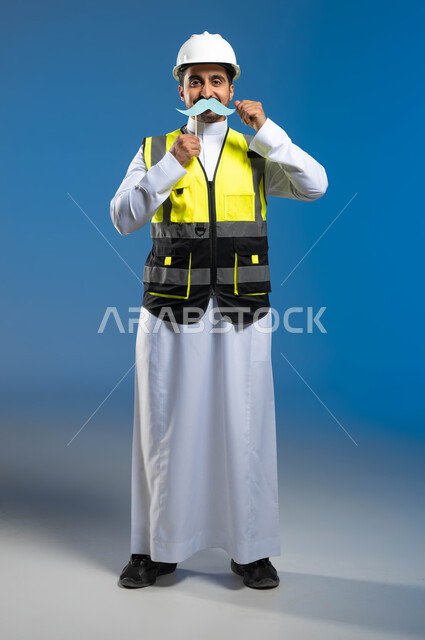 الإحتفال السنوي باليوم العالمي للرجل، العمل في مجال الهندسة والانشاءات المعمارية، بورتريه لمهندس عربي خليجي سعودي يرتدي السترة وخوذة الحماية ويمسك عود خشبي مع قصاصة ورقية على شكل شارب، ايماءات الحفاظ على القوة والصحة الجسدية، الطول الكامل للجسم، خلفية زرقاء